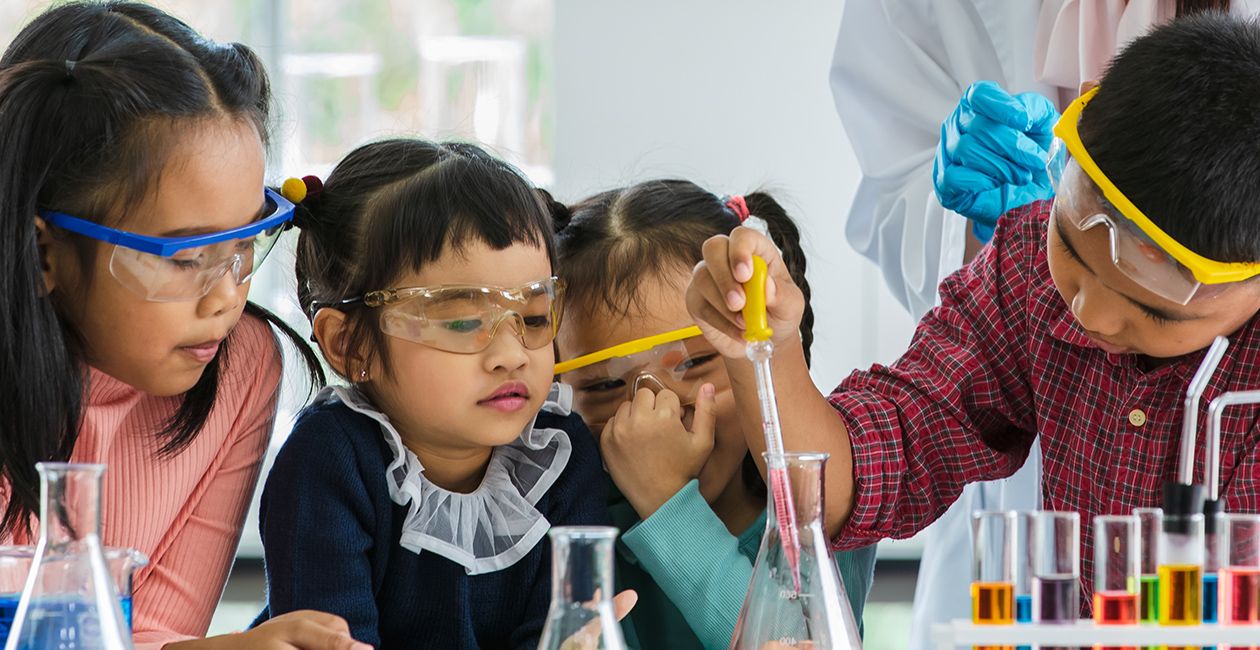 Boy drops something into orange beaker using a pipette in a lab while 3 little girls watch all wearing safety goggles